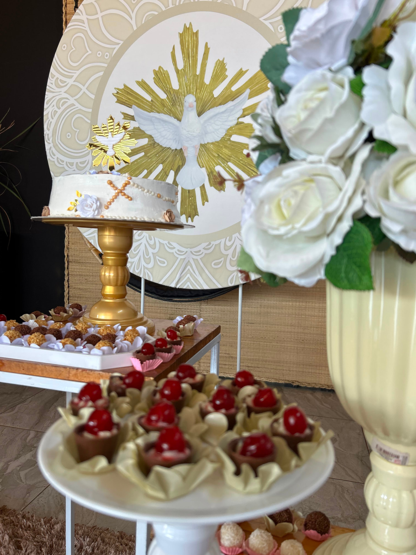 Mesa de doces de batizado com bolo em suporte dourado e painel do Espírito Santo
