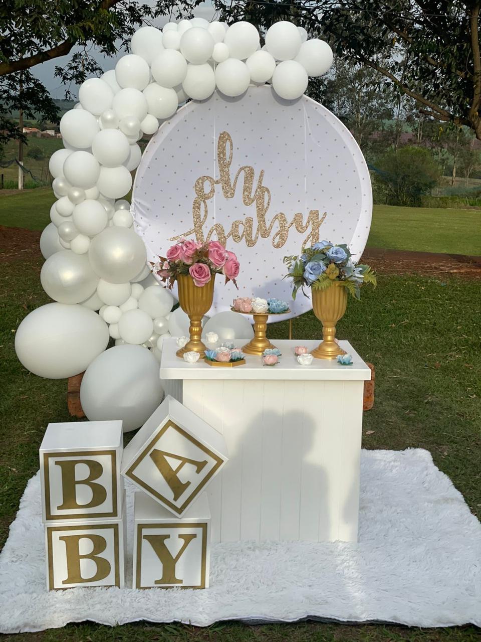 Chá de bebê ao ar livre com painel redondo Oh Baby dourado, mesa branca, vasos gregos com flores e arco de balões brancos