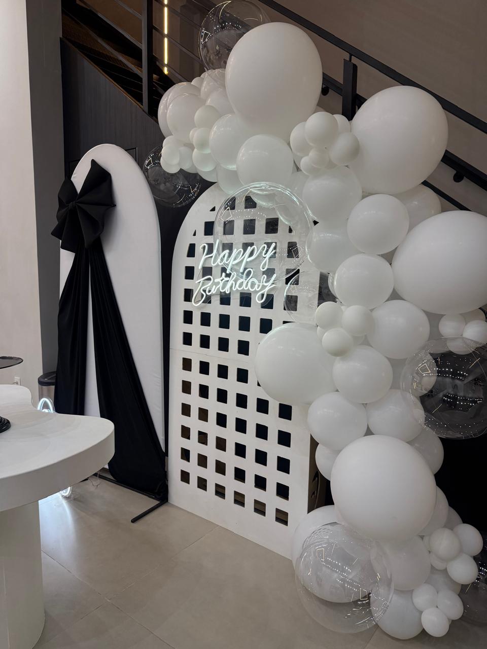 Decoração de aniversário preto e branco com painel vazado, laço preto, letreiro neon Happy Birthday e arco orgânico de balões brancos e transparentes