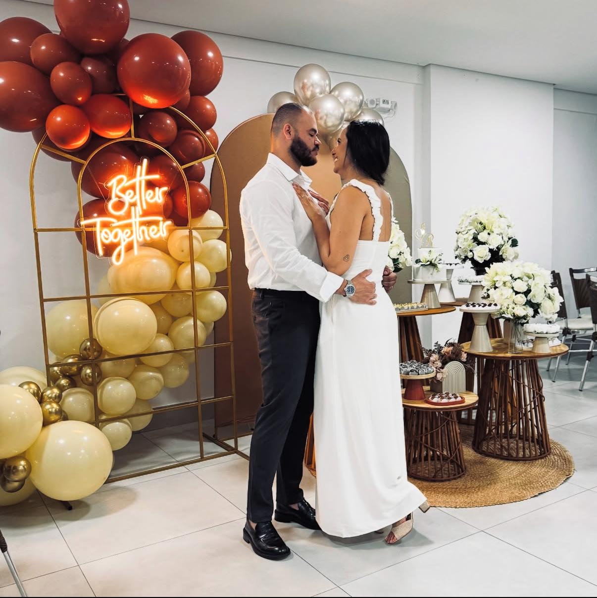 Casal em decoração de casamento com neon Better Together, arco de balões terracota e dourado e mesas de doces