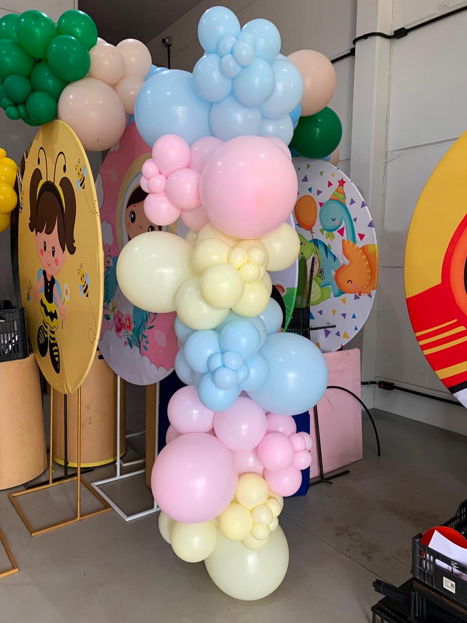 Coluna de balões orgânicos em tons pastel rosa, azul e amarelo com painéis temáticos infantis ao fundo