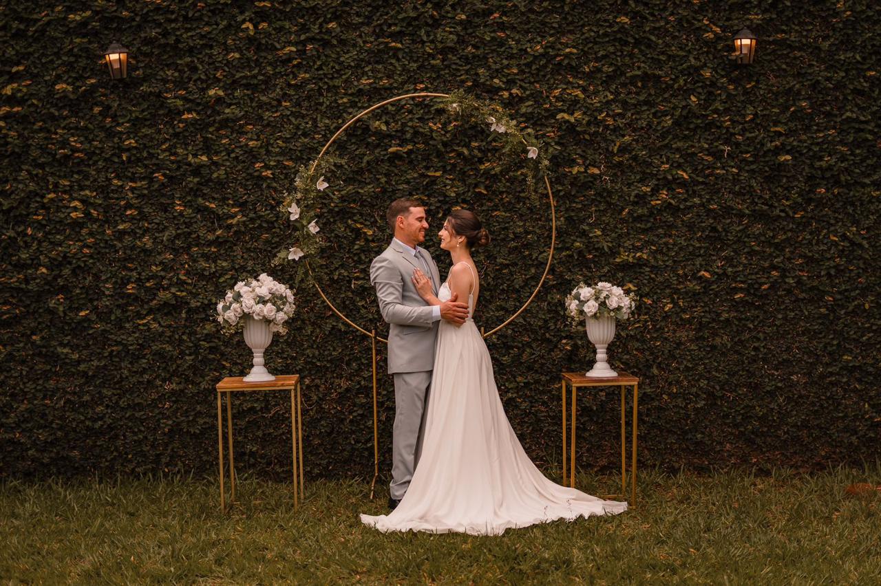 Casamento ao ar livre com arco circular dourado decorado com folhagem e flores, vasos gregos brancos e muro verde ao fundo