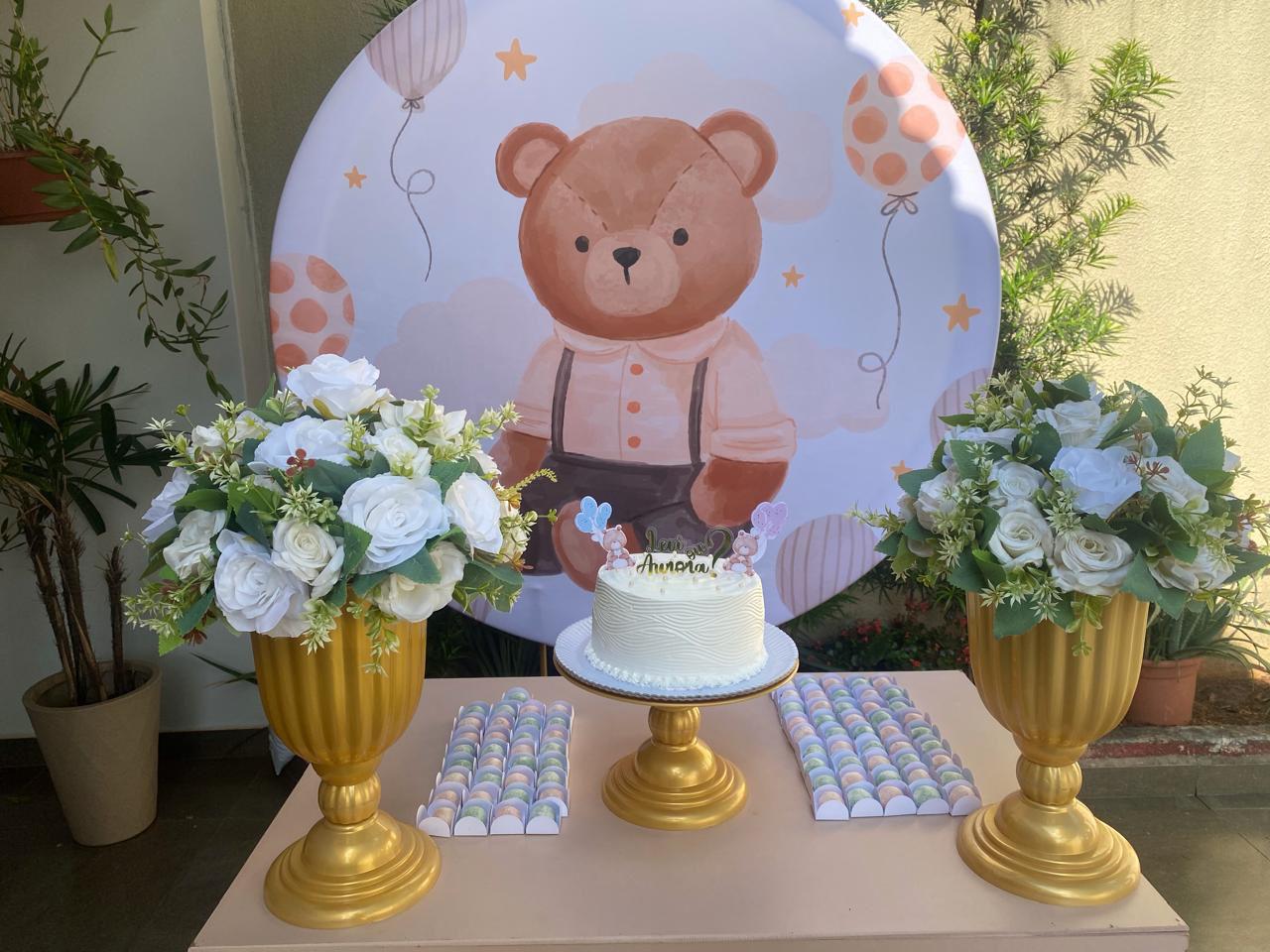Kit montado no tema Ursinho, com painel redondo de urso, bolo branco, arranjos de flores brancas em vasos dourados e docinhos coloridos — decoração delicada para festa infantil